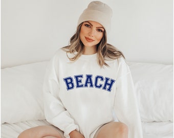 May include: White sweatshirt with the word "BEACH" in navy blue varsity letters. The sweatshirt is worn with a beige beanie. The overall aesthetic is casual and relaxed, perfect for beach lovers.