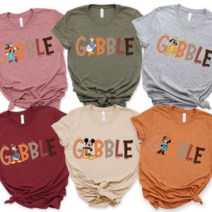 May include: A collection of six t-shirts in various colors, each featuring the word "GOBBLE" in a playful font. Each shirt showcases a different Disney character, including Goofy, Donald Duck, and Mickey Mouse, adding a festive touch.