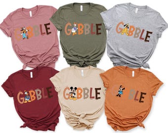 May include: A collection of six t-shirts in various colors, each featuring the word "GOBBLE" in a playful font. Each shirt showcases a different Disney character, including Goofy, Donald Duck, and Mickey Mouse, adding a festive touch.