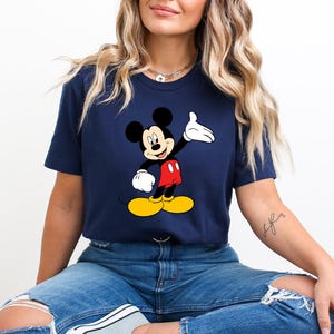 Puede incluir: Camiseta azul marino con un dise&ntilde;o de Mickey Mouse. El personaje de dibujos animados se representa con orejas negras, pantalones cortos rojos, zapatos amarillos y guantes blancos. La camiseta es de cuello redondo.