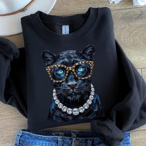May include: A black long-sleeve sweatshirt featuring a graphic print of a black panther wearing leopard-print glasses and a chunky crystal-style necklace. The panther has bright blue eyes and a detailed, textured fur appearance on the front.