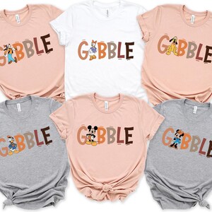 May include: A collection of six short-sleeved t-shirts in various colors, including pink, white, and gray. Each shirt features the word "GOBBLE" in brown, orange, and gray letters, with a different Disney character incorporated into the design.