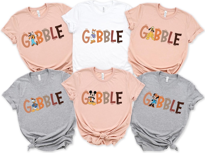 May include: A collection of six short-sleeved t-shirts in various colors, including pink, white, and gray. Each shirt features the word "GOBBLE" in brown, orange, and gray letters, with a different Disney character incorporated into the design.