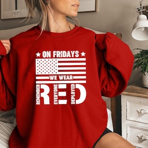 May include: A red sweatshirt with white text and a graphic of the American flag. The text reads "On Fridays We Wear Red" with the words "Remember Everyone Deployed" vertically on the left and right sides.