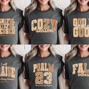 May include: A collection of six dark gray t-shirts with fall-themed text designs. The shirts feature phrases like "Thankful, Grateful, Blessed," "Cozy Season," and "Fall Vibes." The text uses various patterns and colors, including orange, brown, and white.