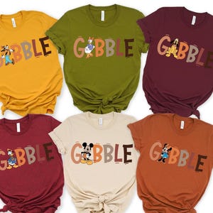 May include: Six colorful t-shirts in mustard yellow, olive green, burgundy, beige, rust, and maroon. Each shirt features the word "GOBBLE" in a fall-themed font, with a different cartoon character illustration.