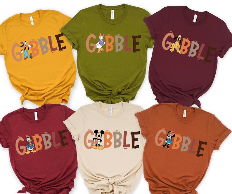 May include: Six colorful t-shirts in mustard yellow, olive green, burgundy, beige, rust, and maroon. Each shirt features the word "GOBBLE" in a fall-themed font, with a different cartoon character illustration.