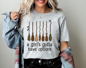 May include: A light gray t-shirt featuring a design of seven different broomsticks in shades of brown and green. The text "a girl's gotta have options" is printed below the broomsticks in black.