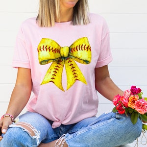 K&ouml;nnte beinhalten: Ein pinkfarbenes T-Shirt mit einem gelben und roten Softball-Schleifendesign. Die Schleife ist so gestaltet, dass sie einem Softball mit roten N&auml;hten &auml;hnelt.