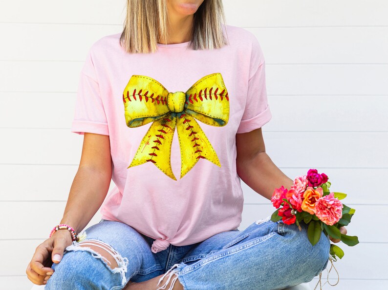 K&ouml;nnte beinhalten: Ein pinkfarbenes T-Shirt mit einem gelben und roten Softball-Schleifendesign. Die Schleife ist so gestaltet, dass sie einem Softball mit roten N&auml;hten &auml;hnelt.