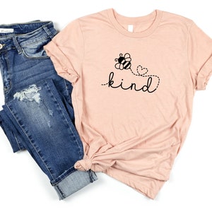 May include: A light pink t-shirt with a black bee and flower graphic and the text "be kind" in a cursive font.