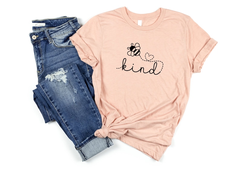 May include: A light pink t-shirt with a black bee and flower graphic and the text "be kind" in a cursive font.