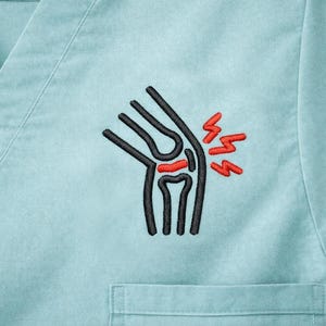 May include: A light blue medical scrub top with a V-neck and a small pocket. The top features an embroidered design of a knee joint with red pain bolts, indicating a medical or healthcare theme.