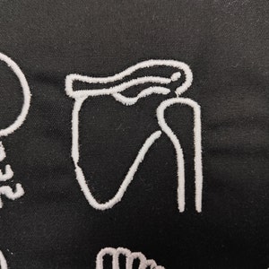Op de afbeelding: Zwarte stof met witte geborduurde anatomische illustraties van botten, waaronder een schoudergewricht. De gedetailleerde borduursels creëren een textuureffect tegen de donkere achtergrond. Het ontwerp is waarschijnlijk voor educatieve of medische doeleinden.