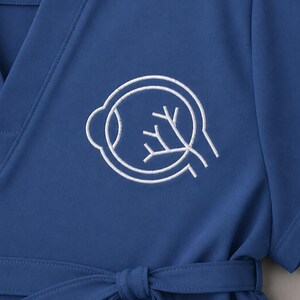 May include: A blue scrub top with a V-neck and a white embroidered eye diagram. The eye diagram features a stylized outline of an eye with internal branching lines. The top has a tie belt at the waist.