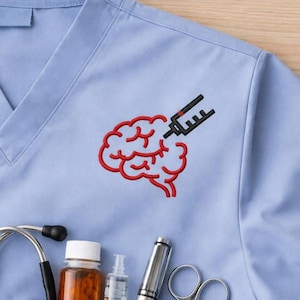 May include: Pale blue medical scrub top with a V-neck and a red brain and syringe embroidered design. A stethoscope, medicine bottles, a syringe, and scissors are visible.