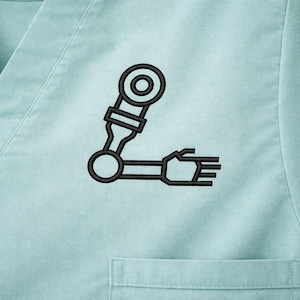 May include: Light blue scrub top with a black embroidered robotic arm design on the chest. The top has a V-neck and a small pocket. The robotic arm is stylized with circular and rectangular shapes.