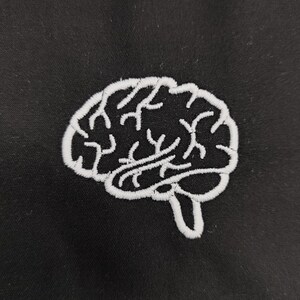 May include: A white embroidered brain design on black fabric. The brain is detailed with lobes and a stem. The stitching is clean and the design is centred. The image is a close-up of the embroidered patch.