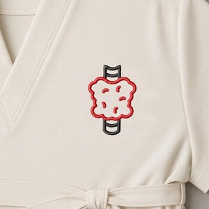 May include: Cream-coloured medical scrub top with a V-neck and a tie at the waist. The top features an embroidered anatomical illustration of a thyroid gland in red, black, and cream colours. The design is centered on the chest.