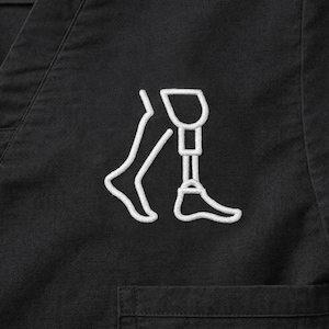 May include: Black scrub top with a white embroidered outline of a leg with a prosthetic leg. The design is located on the upper left chest area of the scrub top. The scrub top has a V-neck and a pocket.