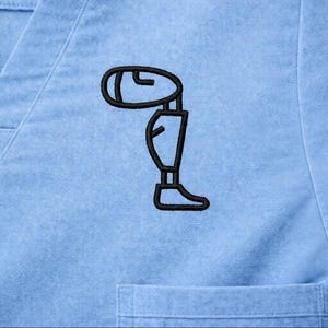 May include: Light blue scrub top with a black embroidered graphic of a prosthetic leg. The top has a V-neck and a small pocket. The graphic is located on the left side of the chest.