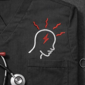May include: Black medical scrub top with a V-neck and a pocket. The top features an embroidered design of a head with a lightning bolt and radiating lines in red and white. A stethoscope and scissors are visible.
