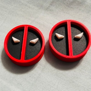 May include: Two red and black circular objects with white eye shapes, resembling the Deadpool logo. The objects have a red border and a black center, with a vertical red line dividing the black area. The white eye shapes are triangular.