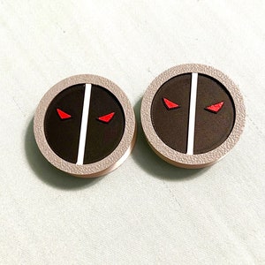 May include: Two round black and white buttons with a red eye design. The buttons are textured and have a white border.