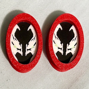 May include: Two red oval-shaped earrings with a white and black image of a character with glowing green eyes. The character's face is centered in the earrings.