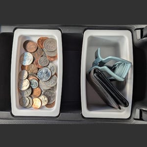 May include: A car console with two white rectangular compartments. One is filled with various coins, the other holds a black wallet and a blue car visor clip. The console is black.