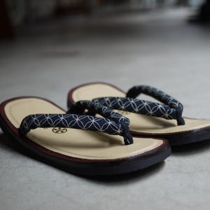 May include: A pair of zori sandals featuring a cream-colored footbed with a dark red trim and a navy blue fabric strap adorned with a white geometric circular pattern. The sandals have a flat, dark sole and a thong-style construction.