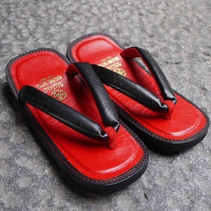 May include: A pair of traditional Japanese-style sandals featuring a vibrant red textured footbed and thick black straps. The heel area displays a gold logo and the text NAGOMO SETTA. The base of the sandals is black with a stitched edge detail.
