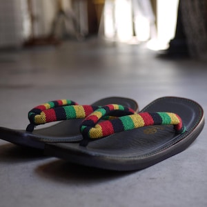 May include: A pair of black sandals featuring thick, rounded straps with a horizontal striped pattern in red, yellow, and green. The footbeds are black with a smooth, contoured surface and a subtle circular logo embossed near the heel area.