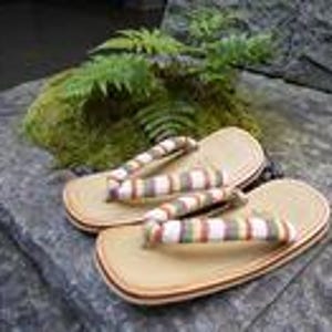 May include: A pair of zori sandals featuring a light tan sole and a thong strap decorated with a multi-colored striped pattern in shades of green, brown, and white. The sandals have a flat, oval-shaped base with a layered edge construction.