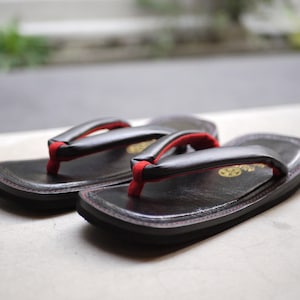 May include: A pair of black zori sandals featuring a smooth black footbed with red stitching along the edges. The thong straps are black with a vibrant red underside and a red fabric toe post. A gold circular emblem is visible on the heel area.