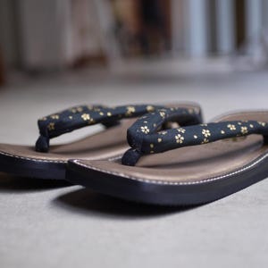 May include: A pair of zori sandals featuring black fabric straps adorned with a small gold floral pattern. The footbeds are a metallic bronze color with a dark brown border, and the soles are black with a thick, slightly curved profile.