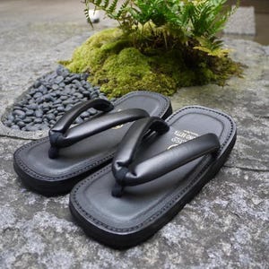 May include: A pair of black setta sandals featuring a flat, rectangular sole with stitched edges and a thick, padded thong strap. The footbed is smooth and dark, with visible branding text printed in gold near the heel area of the sandal.