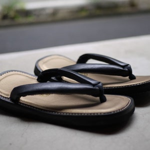 May include: A pair of traditional Japanese-style sandals featuring black padded thong straps and a beige textured footbed. The soles are black with visible stitching along the edges, providing a contrast between the dark exterior and the light interior.