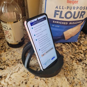 May include: A black phone stand with a smartphone displaying a recipe for banana bread. The recipe instructions are visible on the screen. The phone is on a countertop with a bag of flour and a bottle of vanilla extract in the background.