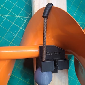 May include: A black 3D printed clamp with a gray ball on the end of a brown cord. The clamp is attached to a wooden surface and is holding a large orange plastic object. The text "Innovations" is printed on the clamp.
