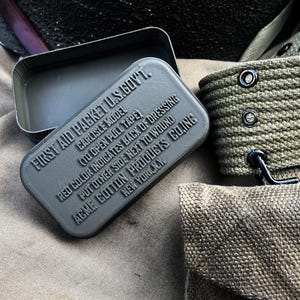 May include: A grey metal first aid kit, open to reveal its interior, with embossed text reading "FIRST AID PACKET U.S.GOVT." and "CARLISLE MODEL." It sits on a tan fabric, with olive-green canvas straps and a pouch visible. The text includes instructions and the manufacturer's location.