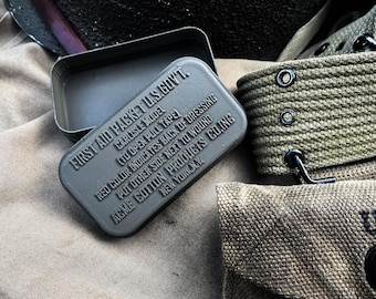 May include: An open, gray metal first aid kit with embossed text, labeled "FIRST AID PACKET U.S.GOVT." and "CARLISLE MODEL." The kit rests on a tan fabric surface, alongside olive-green canvas straps and a pouch. The text includes instructions and the manufacturer's location.