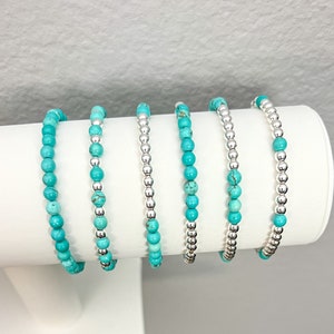 May include: Six turquoise and silver beaded bracelets on a white display stand. The bracelets are made with small, round beads in alternating turquoise and silver colors.