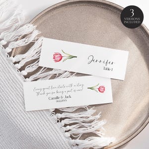 May include: Two white place cards with a watercolor tulip design and text. One card reads "Jennifer, Table 1." The other card has the text "Every great love starts with a story. Thank you for being a part of ours! Camille & Jack 061220XX."
