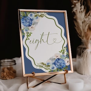 May include: A framed table number with a gold metal stand. The card features a blue background with a white, scalloped border. The word "eight" is written in cursive, surrounded by watercolor flowers.