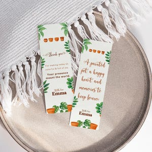 Puede incluir: Dos marcapáginas blancos con ilustraciones botánicas y texto. Uno dice "Gracias" y el otro dice "Una maceta pintada, un corazón feliz y recuerdos para siempre". Ambos incluyen el nombre "Emma".