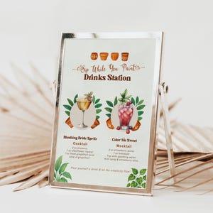May include: A silver framed sign for a drinks station. The sign has the text "Sip While You Paint Drinks Station" at the top. Below are two drink recipes, "Blushing Bride Spritz" and "Color Me Sweet". The sign is propped up by a silver stand.