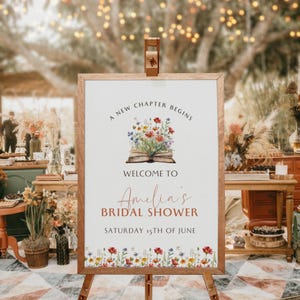 Puede incluir: Un cartel blanco con un marco de madera marrón que anuncia "A new chapter begins" con una ilustración de un libro abierto con flores en la parte superior. El texto "Welcome to Amelia's Bridal Shower Saturday 15th of June" está impreso debajo de la ilustración.