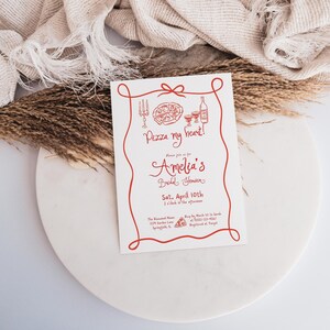 May include: A red and white bridal shower invitation with a pizza and wine illustration. The invitation reads "Pizza my heart!" and includes the text "Please join us for Amelia's Bridal Shower, Sat. April 10th, 2 o'clock in the afternoon, The Reserved Manor, 1234 Garden Lane, Springfield, IL 75501 231-4567, RSVP by March 1st to Sarah, Registered at Target."