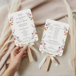 May include: Two wedding fans with floral designs and text. The fans have a wooden handle and a white ribbon. The text on the fans includes the names "Emily Grace and Andrew James" and "Wedding Party".